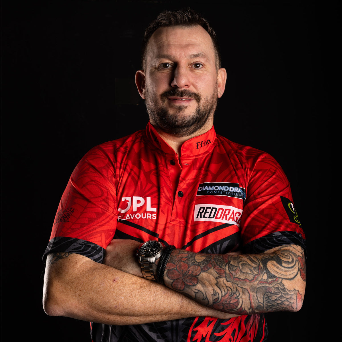 Darts player Jonny Clayton in his sponsored Red Dragon shirt, tattooed arm crossed over the other, black studio background