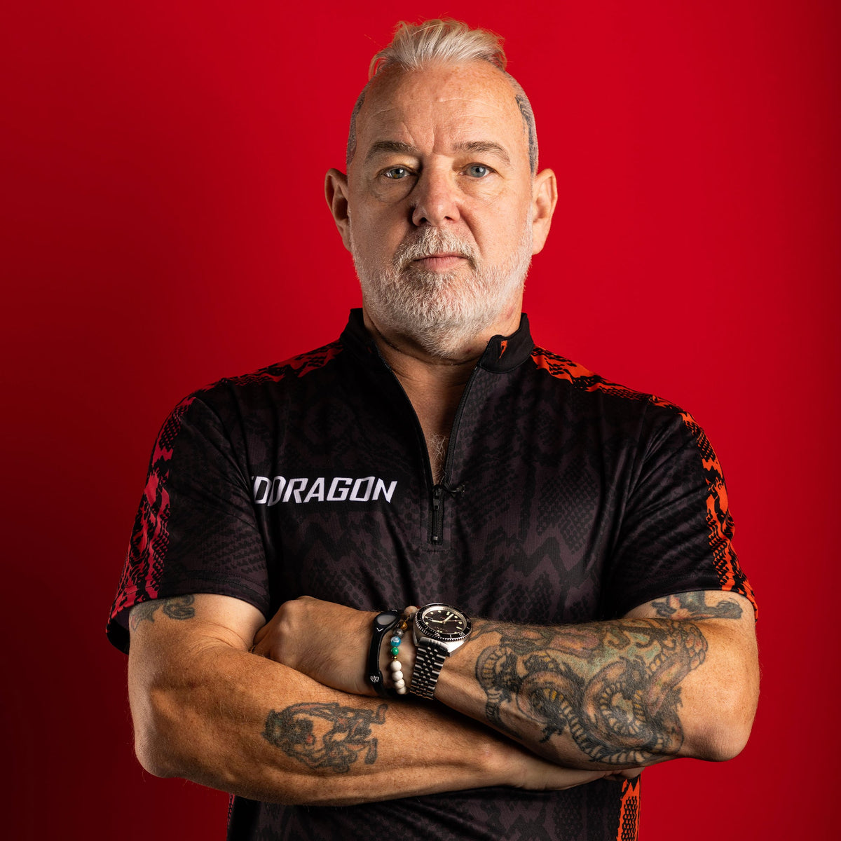 Darts player Peter Wright in his sponsored Red Dragon shirt, arms crossed showing tattooed forearms, red studio background