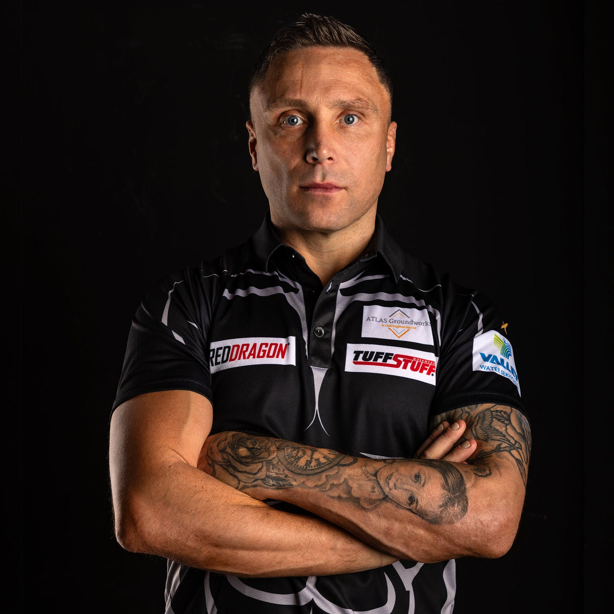 Darts player Gerwyn Price in his sponsored match shirt, arms crossed showing tattooed forearms, black studio background