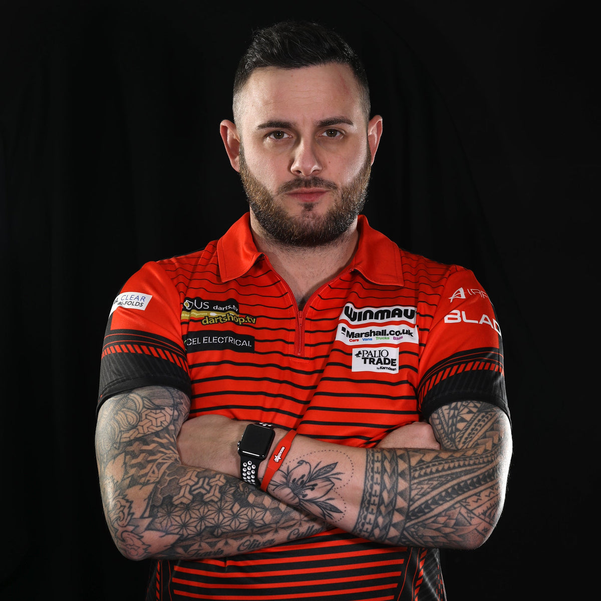 Darts player Joe Cullen in his sponsored Winmau match shirt, tattooed arms crossed, black studio background