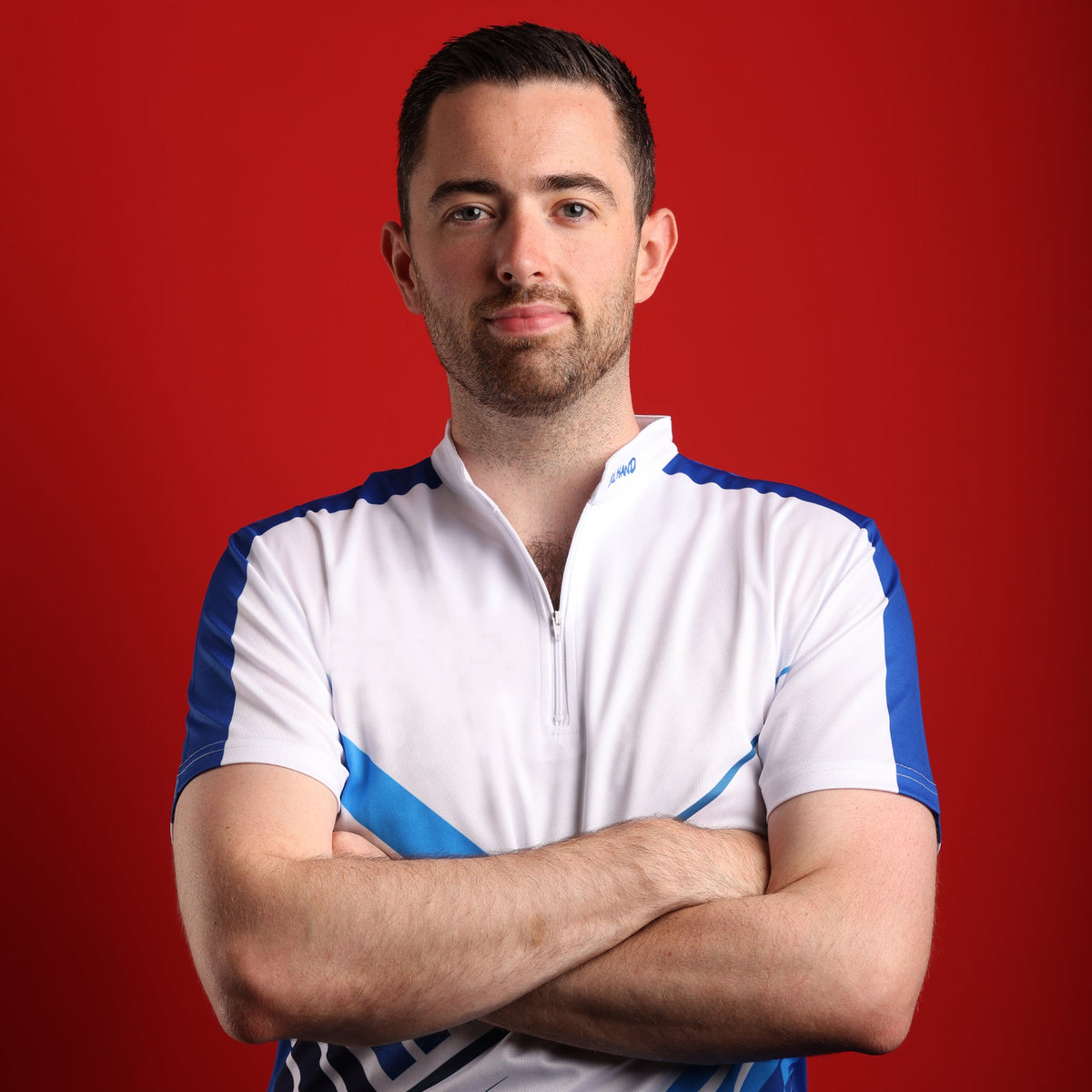 PDC darts player Luke Humphries stood with arms crossed in his tournament shirt, red studio backdrop
